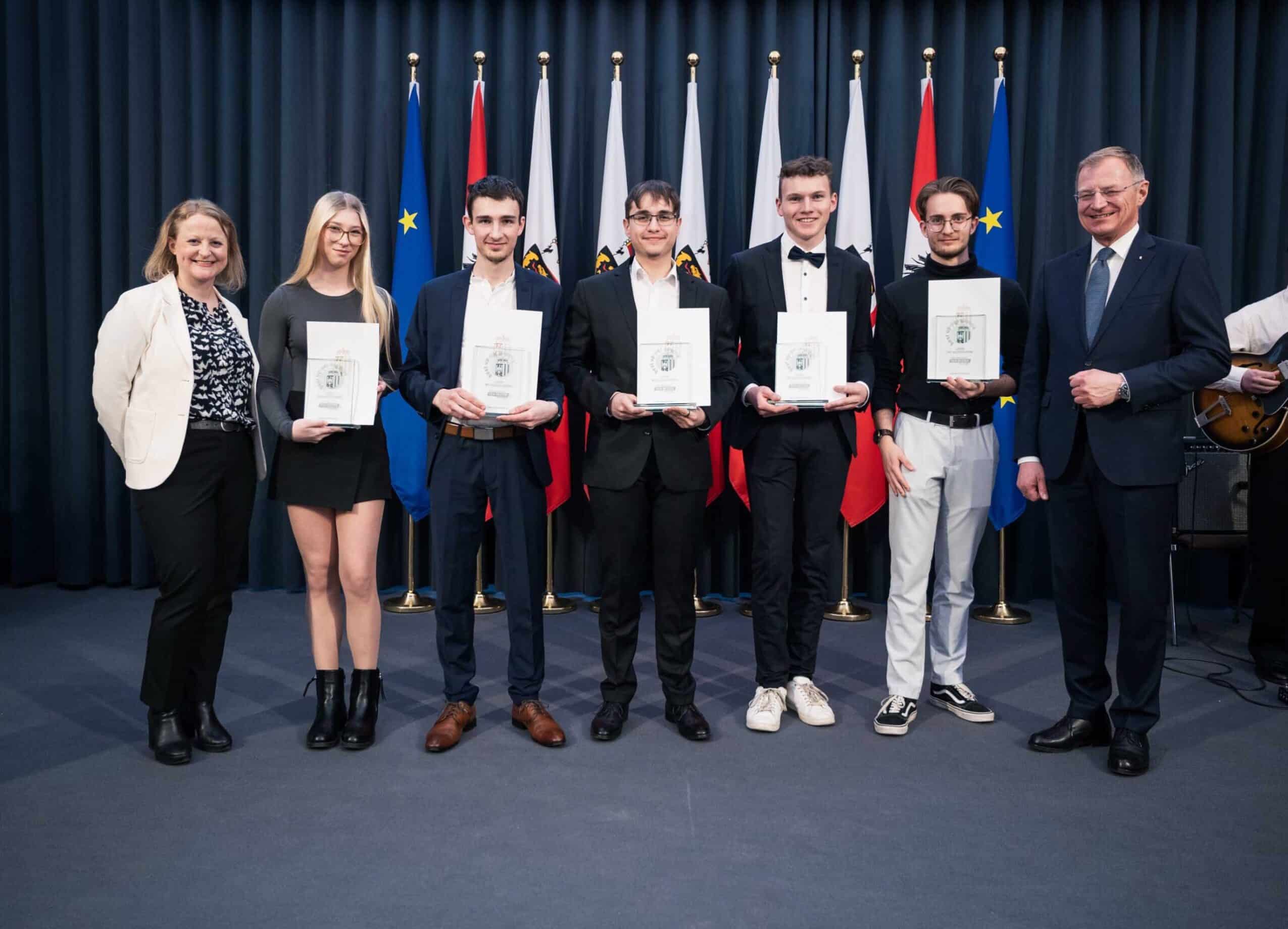 Gruppe von fünf jungen ETECH-Lehrlingen und zwei Ehrengästen bei einer Auszeichnungsfeier mit Urkunden vor Flaggen.