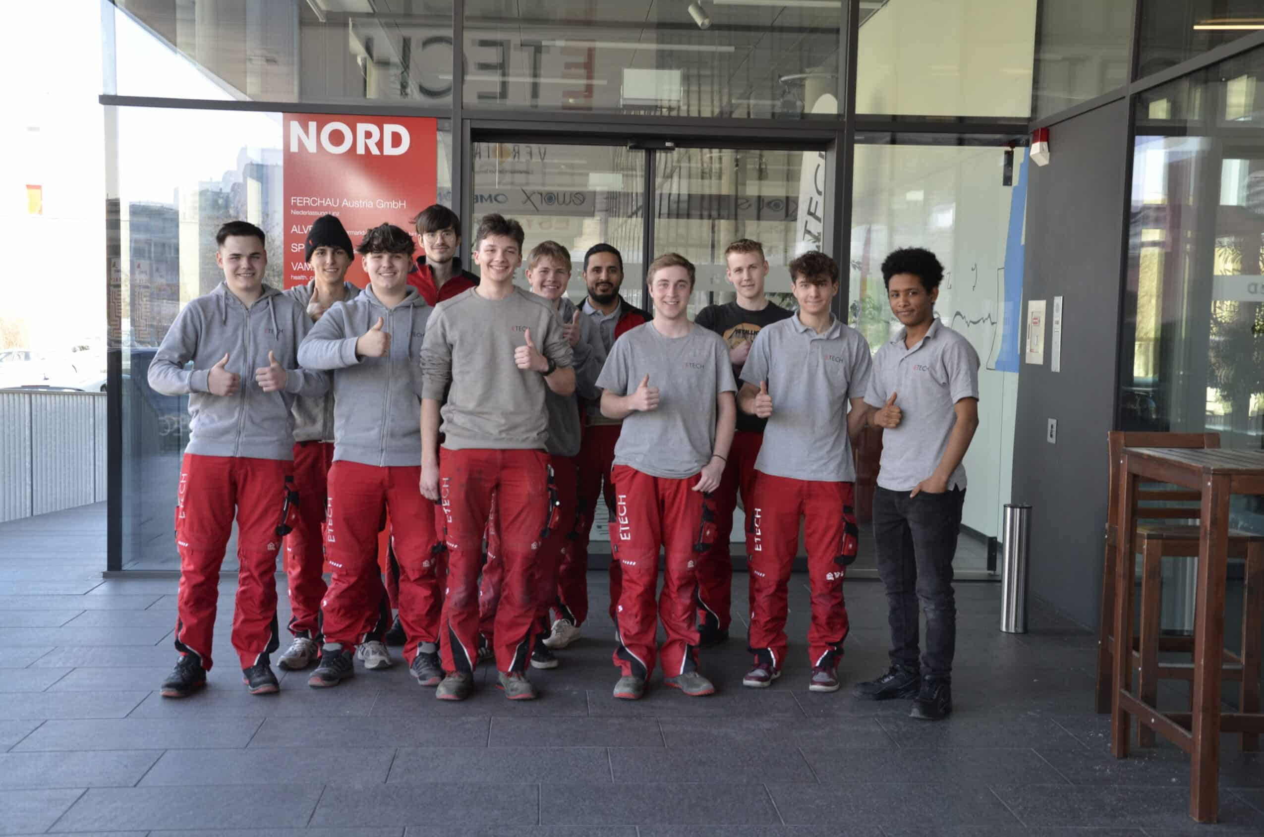 Gruppe von 11 ETECH-Lehrlingen in roter Arbeitskleidung zeigt Daumen hoch vor dem NORD-Gebäude in Linz