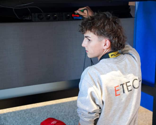 Junger ETECH-Mitarbeiter in grauem Sweatshirt arbeitet an elektrischer Anlage, Fluke-Messgerät um den Hals