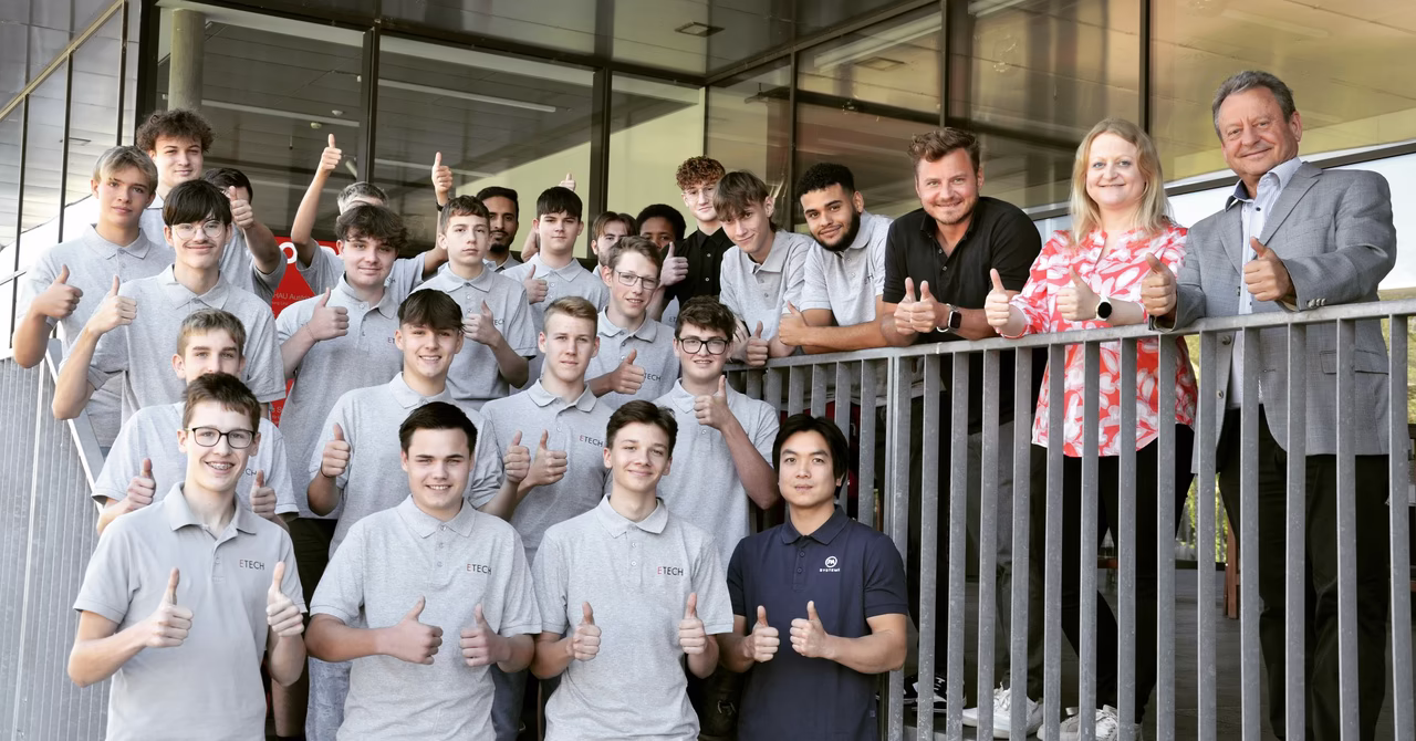 Gruppe von ETECH-Lehrlingen 2023 in grauen Firmen-Poloshirts mit Daumen hoch, begleitet von Klaus und Sophie Schmid – Gruppenfoto vor dem ETECH-Gebäude.