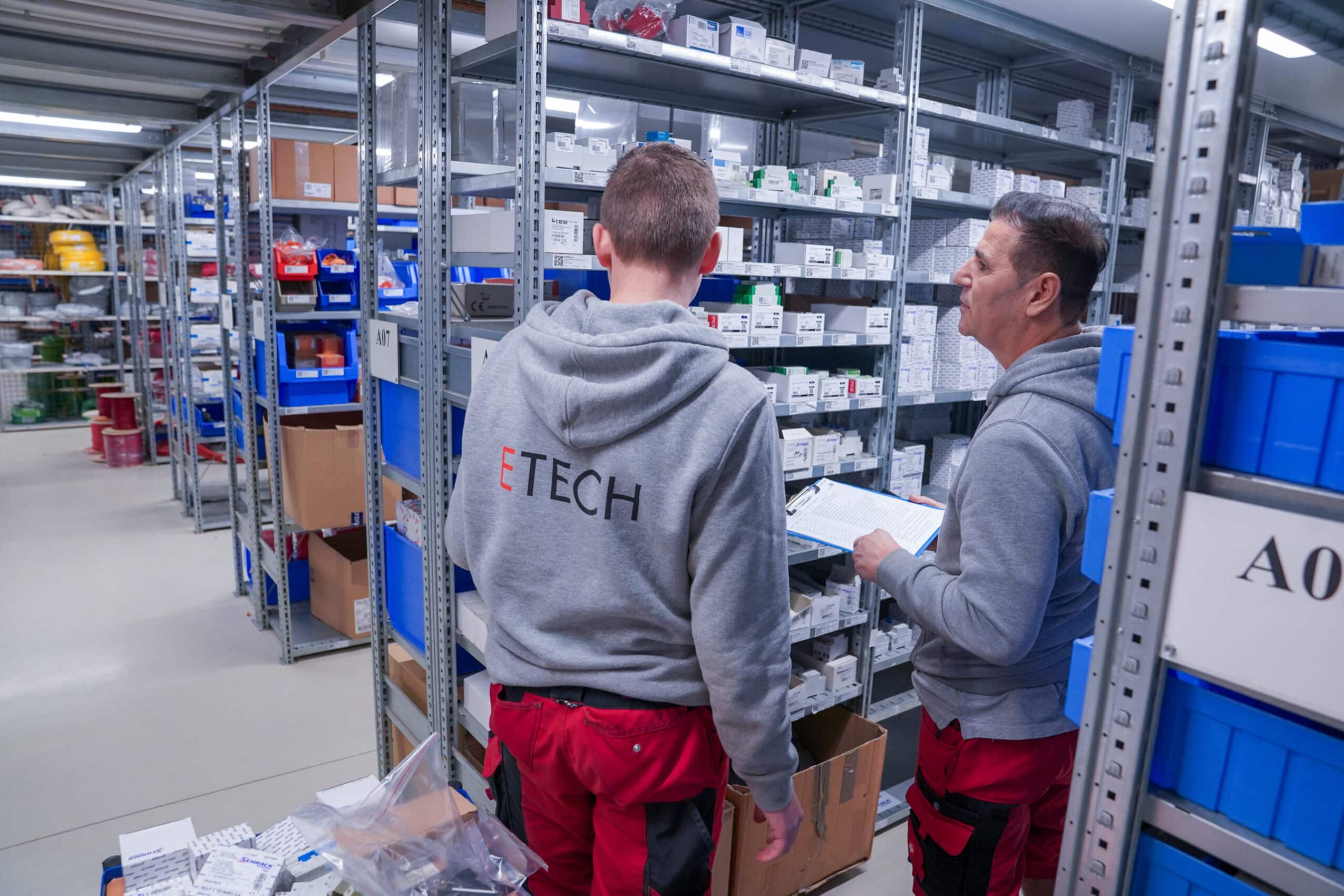 Zwei ETECH-Mitarbeiter in grauer Firmenkleidung und roten Arbeitshosen prüfen Lagerbestände in einem gut sortierten Elektrotechnik-Lager.