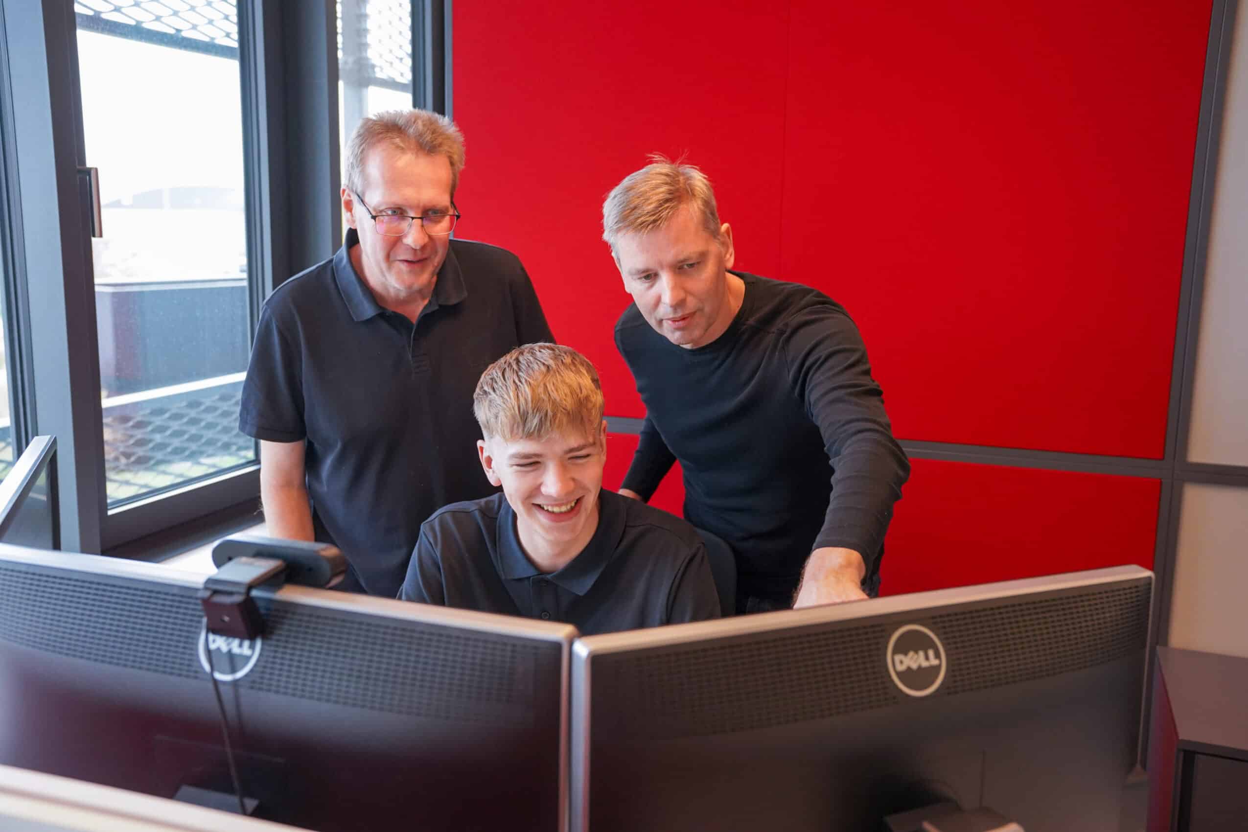 Zwei ETECH-Mitarbeiter zeigen einem lachenden Lehrling etwas an zwei Dell-Monitoren in einem modernen Büro mit roter Wand.