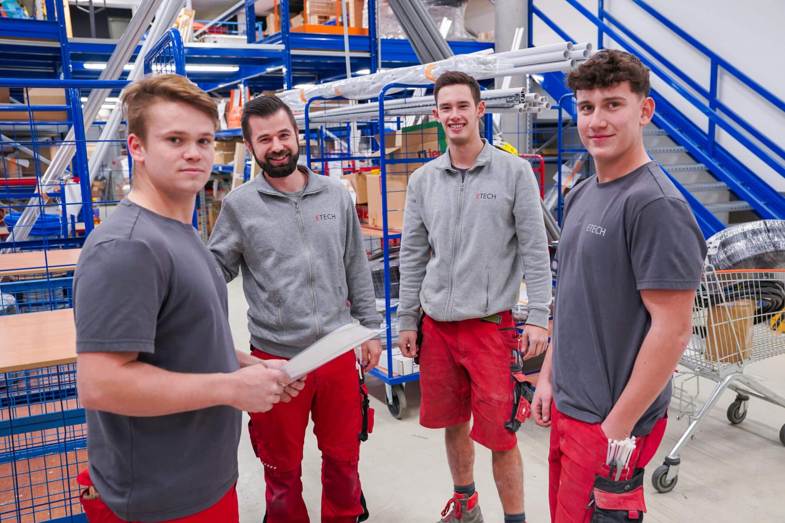 Vier ETECH-Mitarbeiter in grauer und roter Arbeitskleidung in einem Warenlager des Unternehmens ETECH Schmid u. Pachler Elektrotechnik.