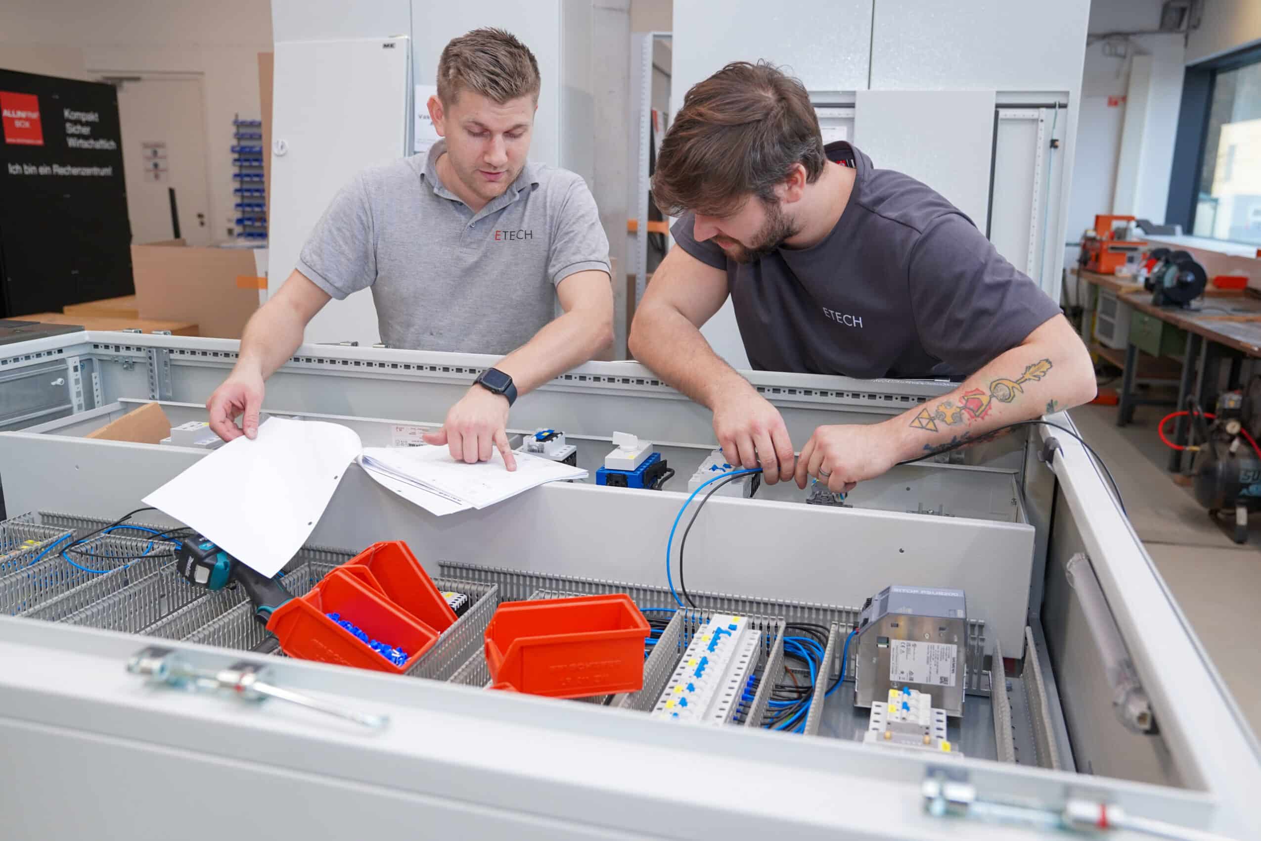 Zwei ETECH-Mitarbeiter arbeiten gemeinsam an einem geöffneten Schaltschrank und prüfen Schaltpläne in einer Werkstatt.