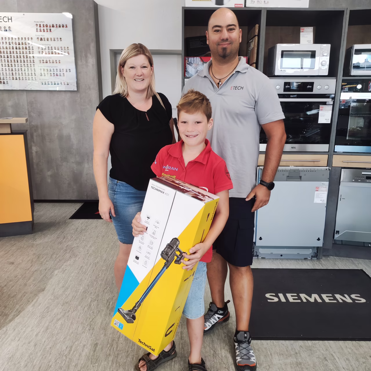 Gewinnerin mit Kind und ETECH-Mitarbeiter hält TechniSat Technimax AS1 Staubsauger im ETECH-Fachhandel
