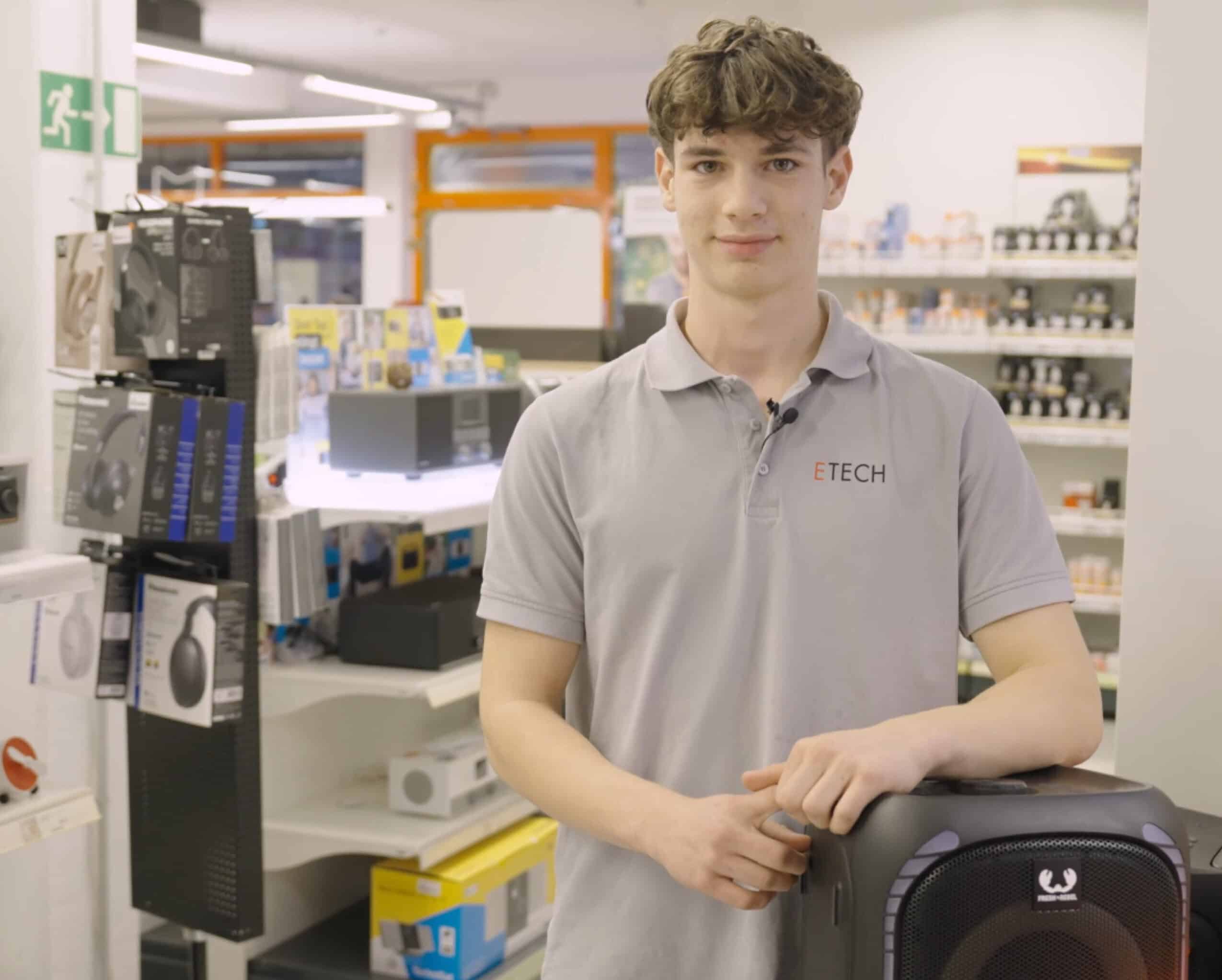 Junger ETECH-Mitarbeiter in grauem Polo-Shirt lehnt an einem Lautsprecher im Elektrofachhandel Gmunden