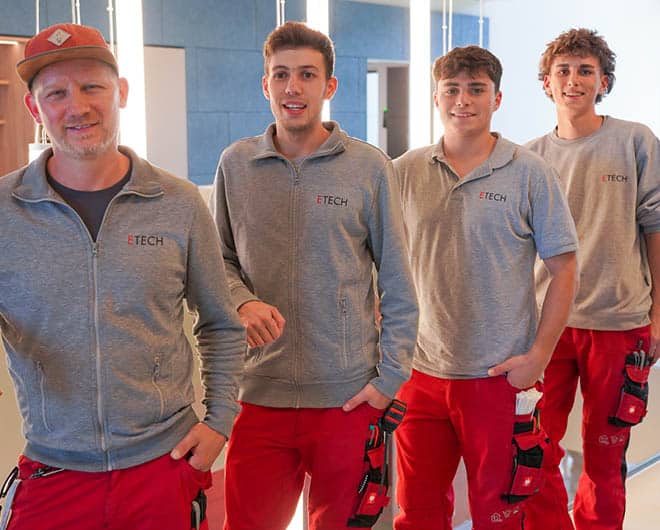 Vier ETECH-Mitarbeiter in grauer Arbeitskleidung und roten Arbeitshosen posieren lächelnd für ein Teamfoto – Einblick in die Lehrausbildung bei ETECH.