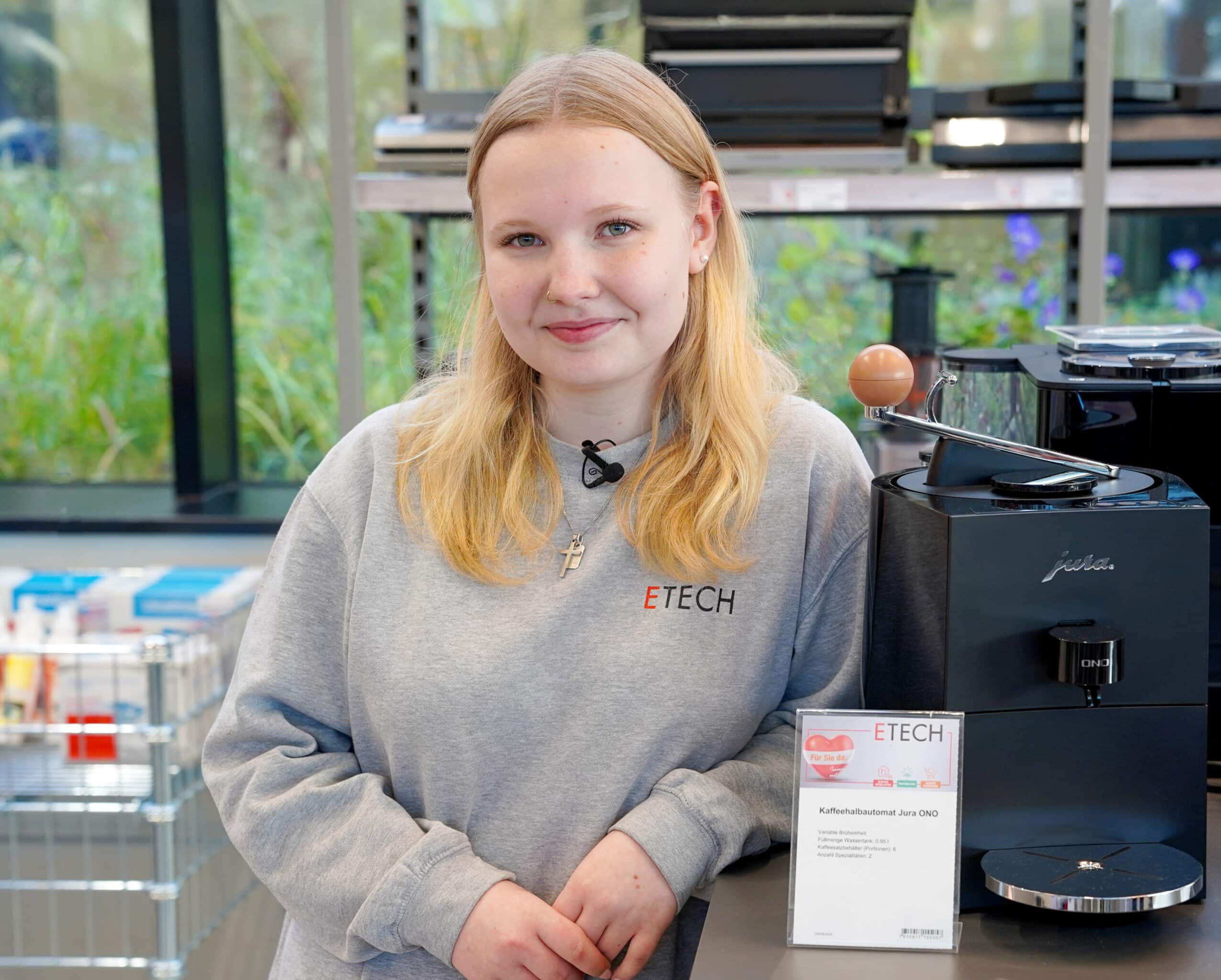 Lehrling bei ETECH Schmid & Pachler mit grauem ETECH-Sweatshirt vor einer Jura ONO Kaffeemaschine im Elektrofachhandel