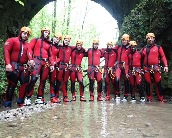 Gruppe von ETECH-Lehrlingen in roten Neoprenanzügen und Helmen beim Canyoning-Ausflug vor einer Felsbrücke