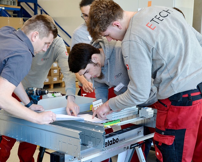 ETECH-Lehrlinge und Ausbilder arbeiten gemeinsam an einem Elektroinstallationsprojekt auf einer Werkbank im Lehrlingworkshop.