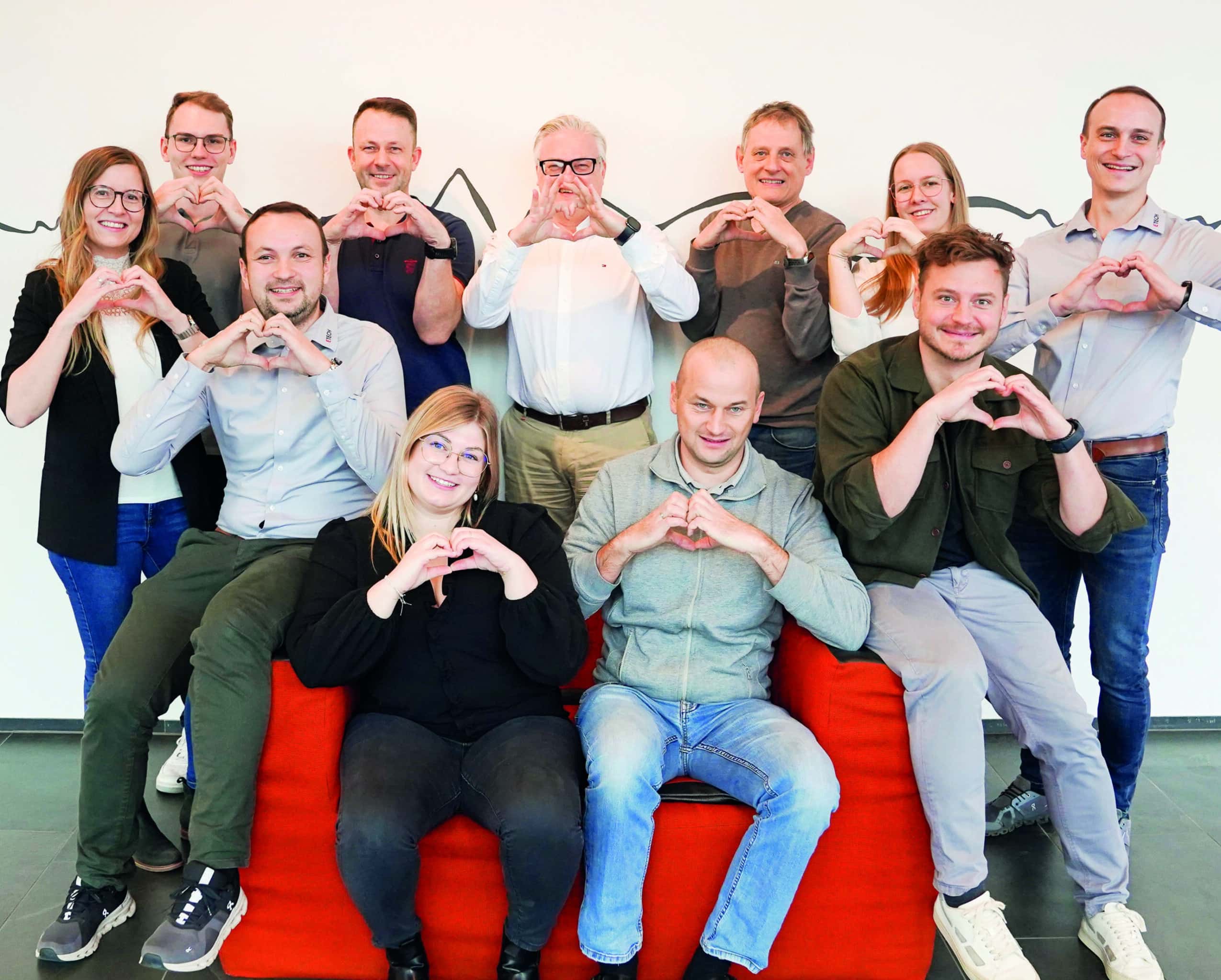 ETECH-Team formt Herzen mit den Händen – Gruppenfoto mit 11 Mitarbeitenden auf rotem Sofa