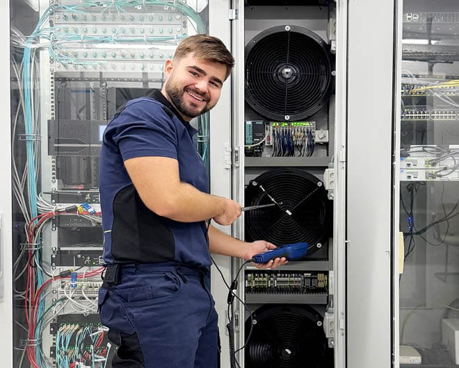 Lächelnder ETECH-Mitarbeiter bei der Wartung eines Schaltschranks mit Lüftern und Verkabelung in einer Elektroanlage
