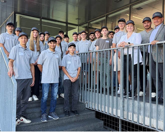 Gruppe von ETECH-Lehrlingen in grauen Poloshirts und TECH-Caps beim Willkommenstag, posierend auf einer Treppe zusammen mit Klaus, Florian und Sophie Schmid