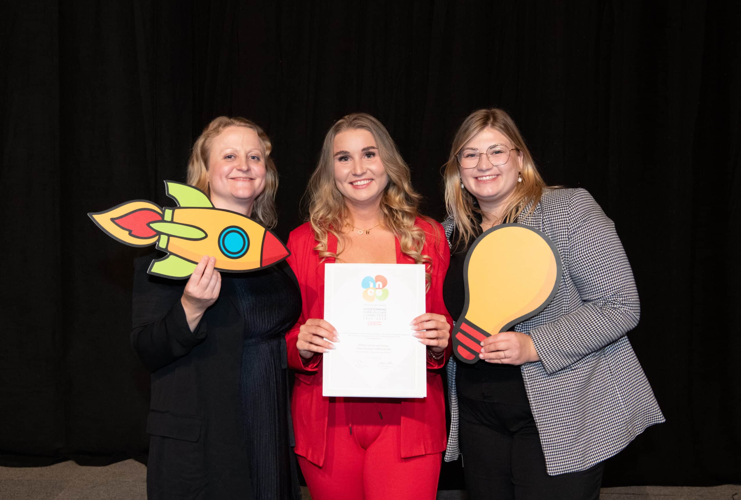 Drei lächelnde Frauen posieren mit einer Urkunde sowie Raketen- und Glühbirnen-Requisiten bei einer Auszeichnungsveranstaltung.